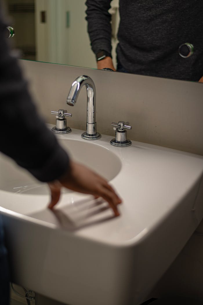 Man's reflection in a bathroom mirror above a ceramic sink with chrome faucet.